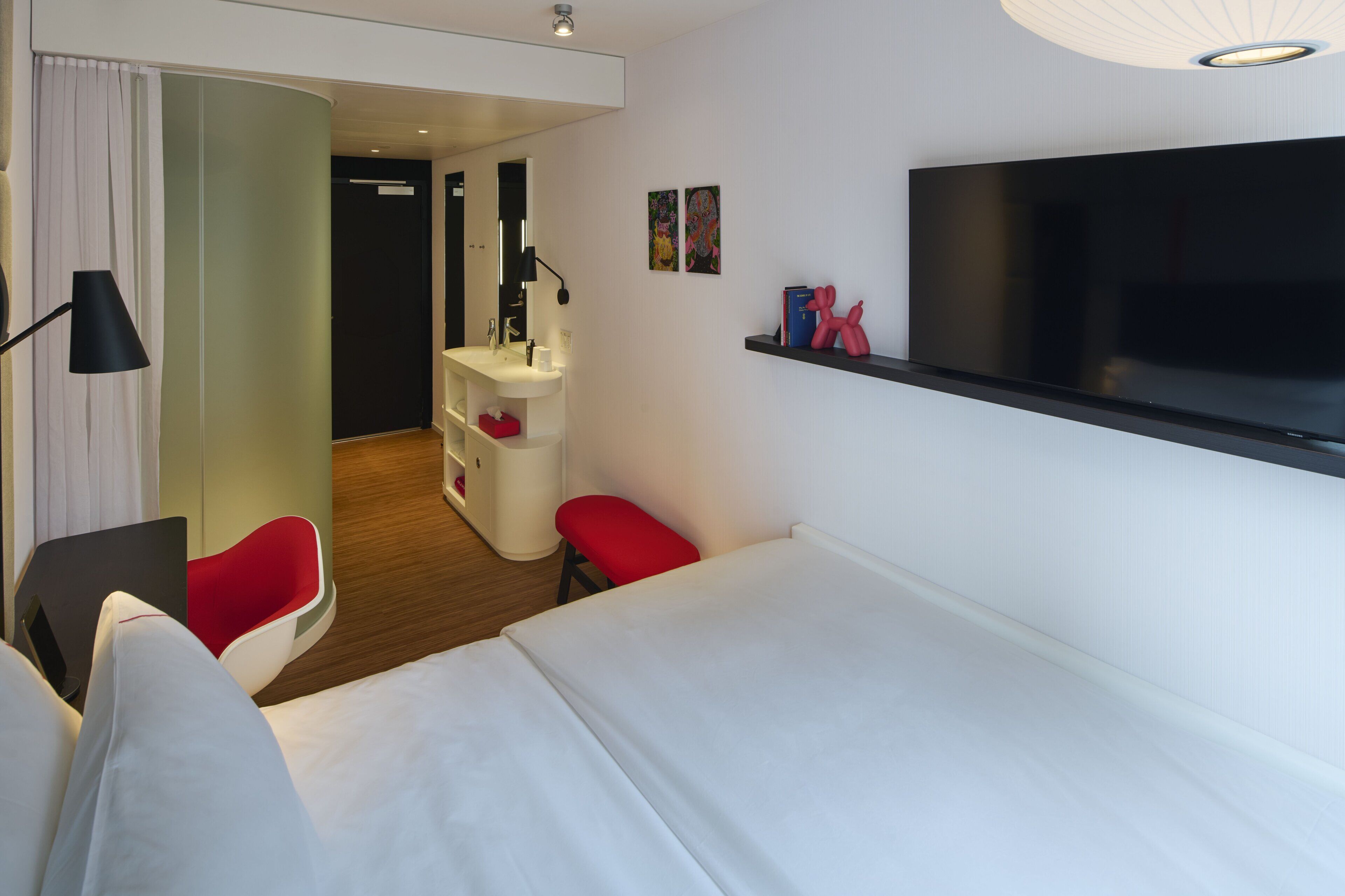 A hotel room with a bed, a desk with a chair, a vanity with a mirror, a television mounted on the wall, and a red bench.