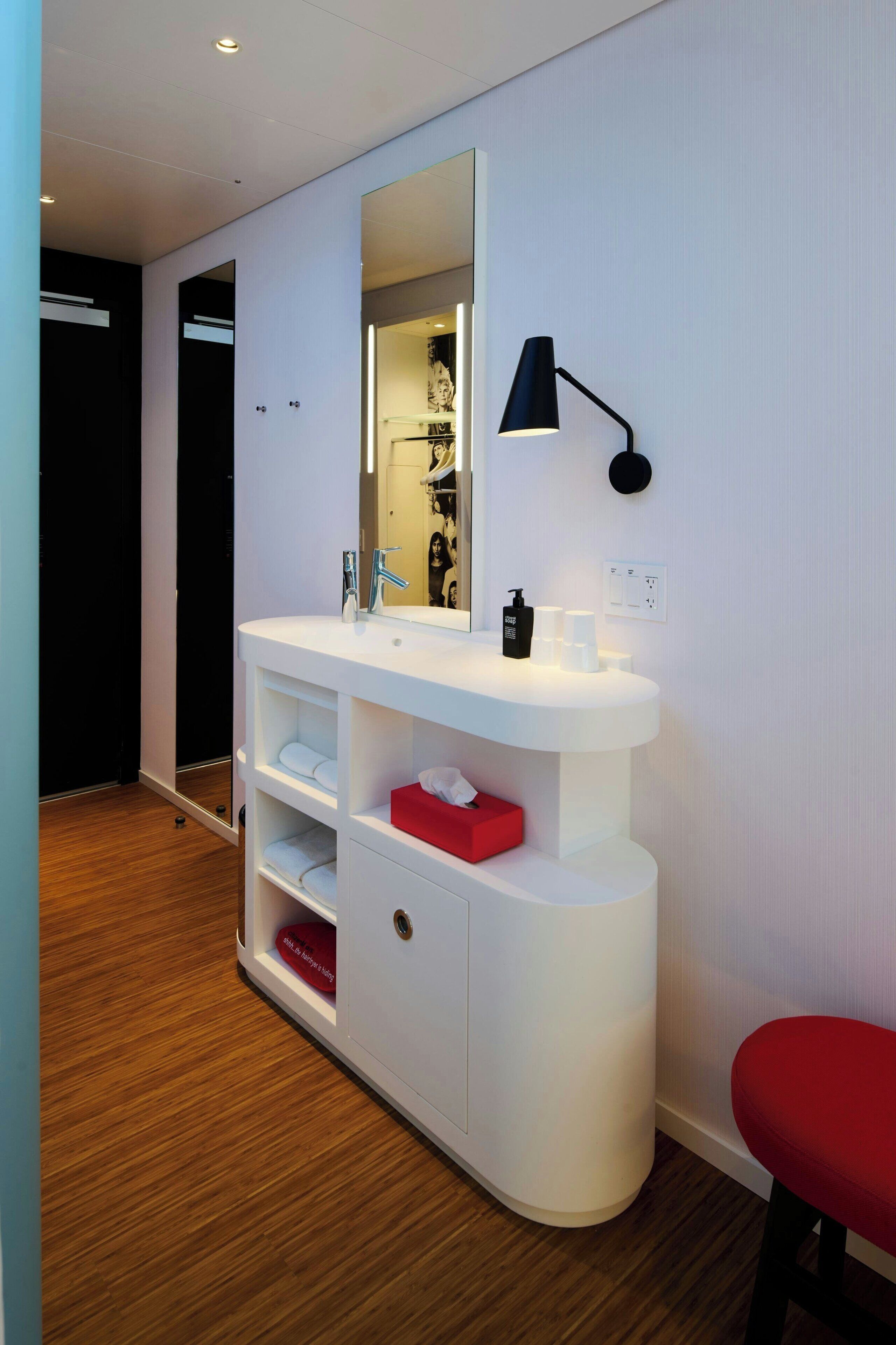 Ein modernes Badezimmer mit einem weißen Waschtisch, einem Spiegel, einer schwarzen Lampe und einem roten Hocker.