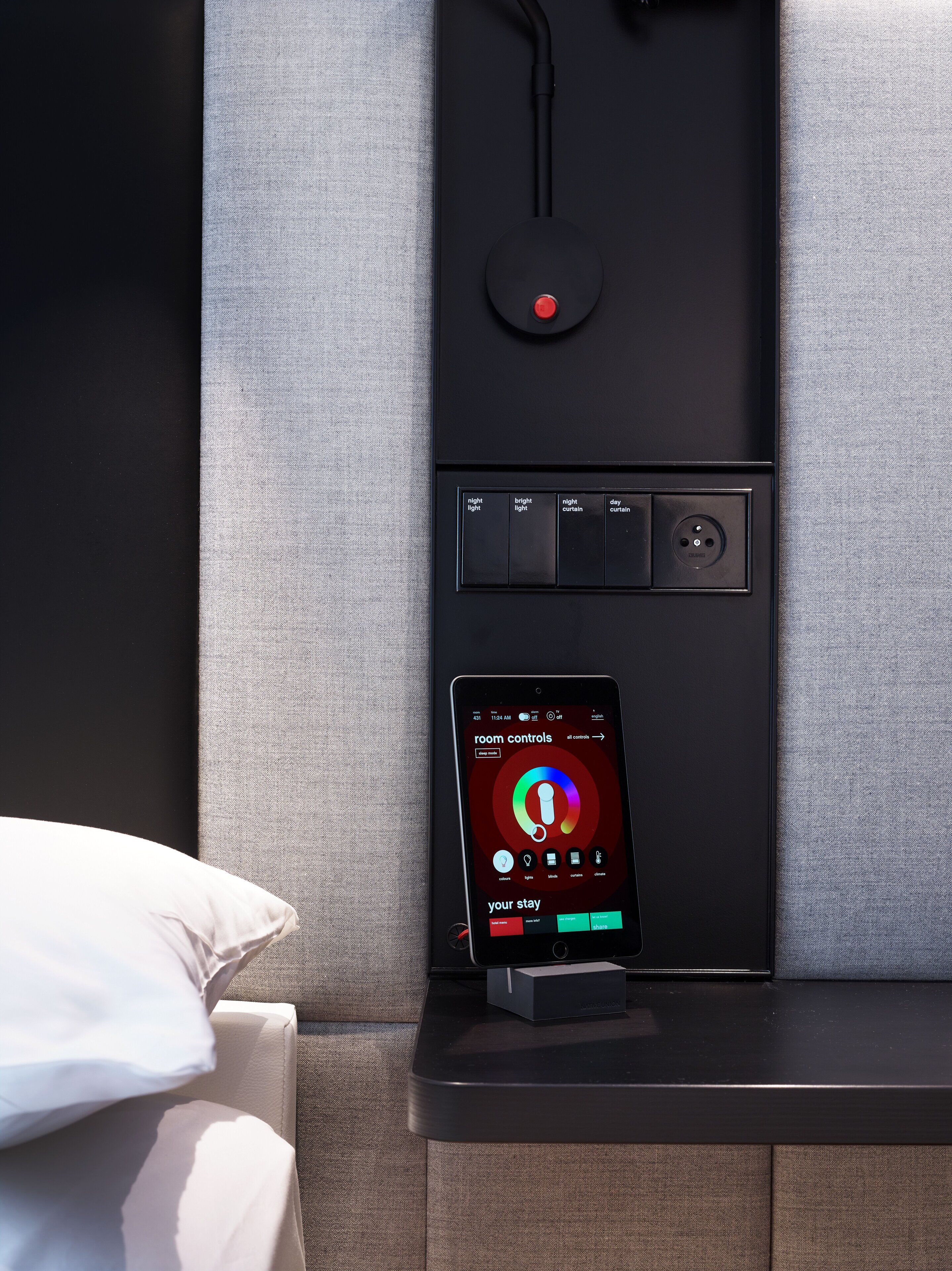 Un panel de control moderno con un teléfono inteligente que muestra los controles de la habitación, un enchufe y un botón rojo.