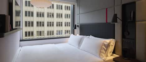 A modern hotel room with a large bed, a bedside table, a lamp, and a window with a city view.