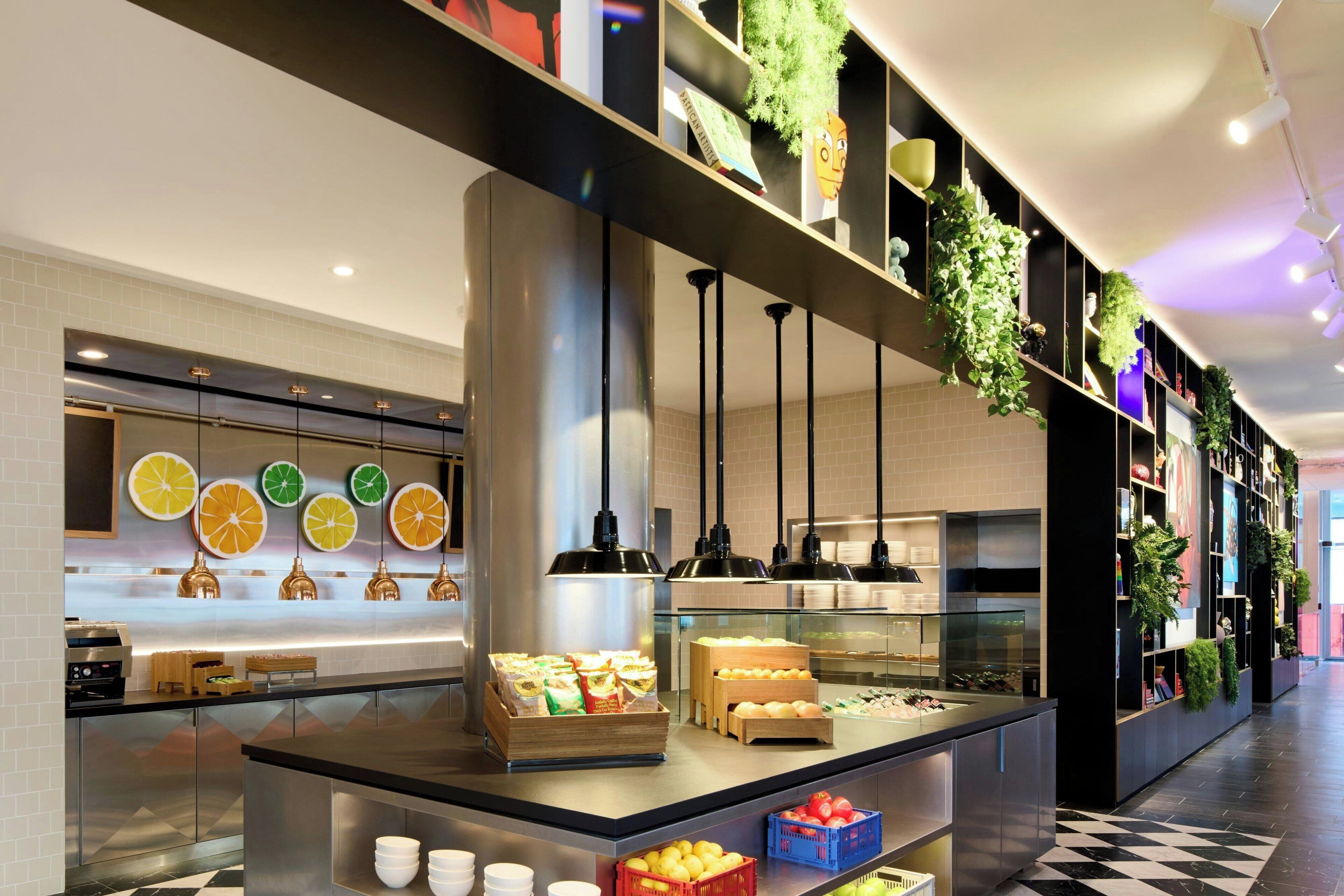 Een moderne hotellobby met een centraal gelegen balie waarop etenswaren staan, een betegelde vloer en een wand versierd met groen en ingelijste foto's.