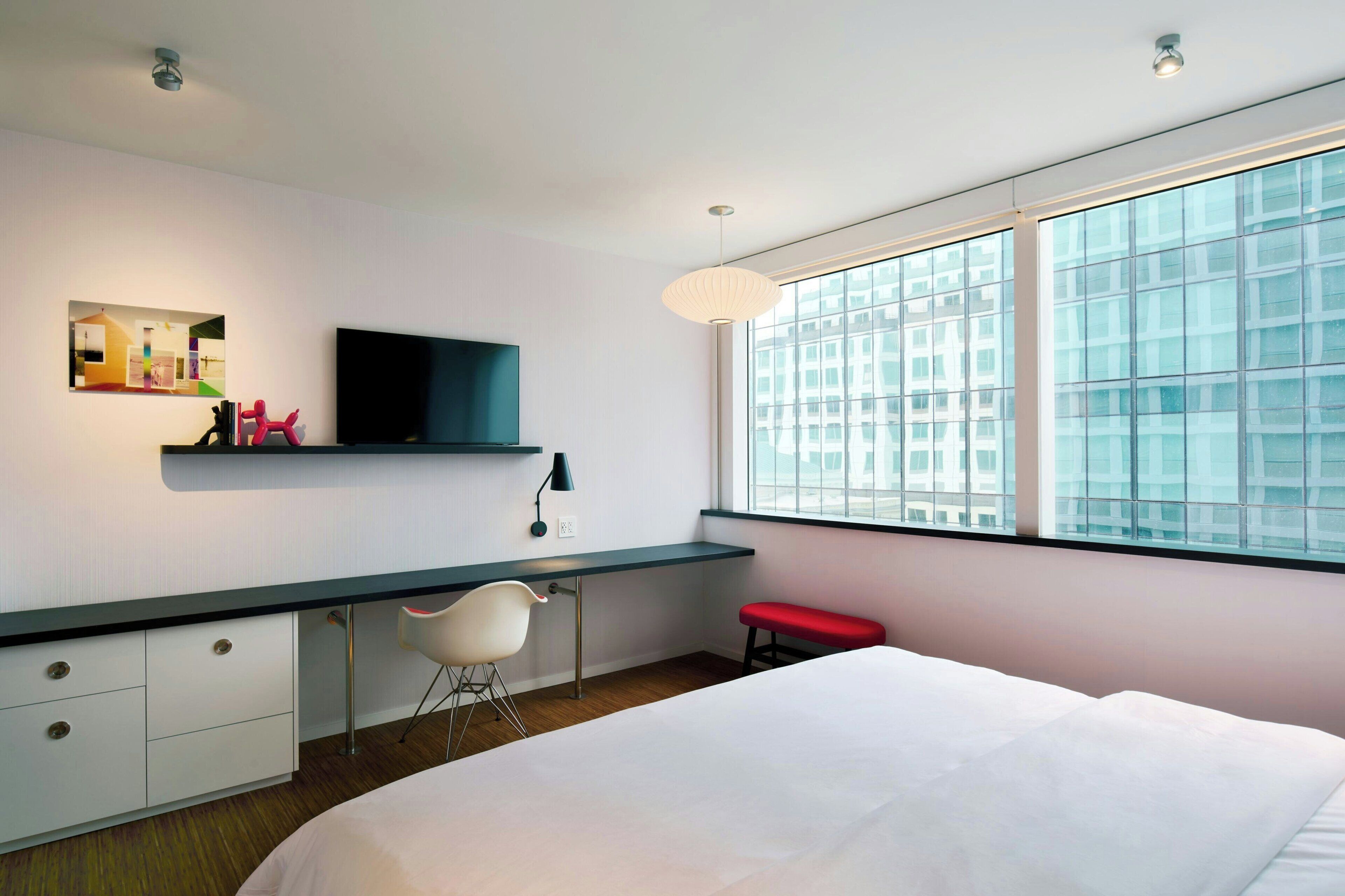 A modern hotel room with a large bed, a desk with a chair, a TV mounted on the wall, and a large window with a city view.