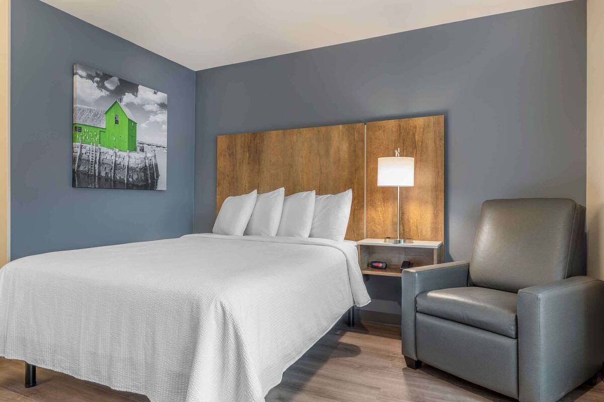Une chambre d’hôtel avec un lit, un fauteuil gris, une tête de lit en bois, une table de chevet et une lampe.