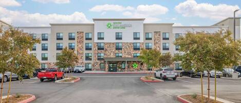 Ein Hotel mit modernem Design, das einen zentralen Eingang mit der Beschilderung 'Extended Stay America Premier Suites' zeigt. Mehrere Parkplätze sind sichtbar, und das Gebäude wird von begrünten Flächen mit jungen Bäumen umgeben.