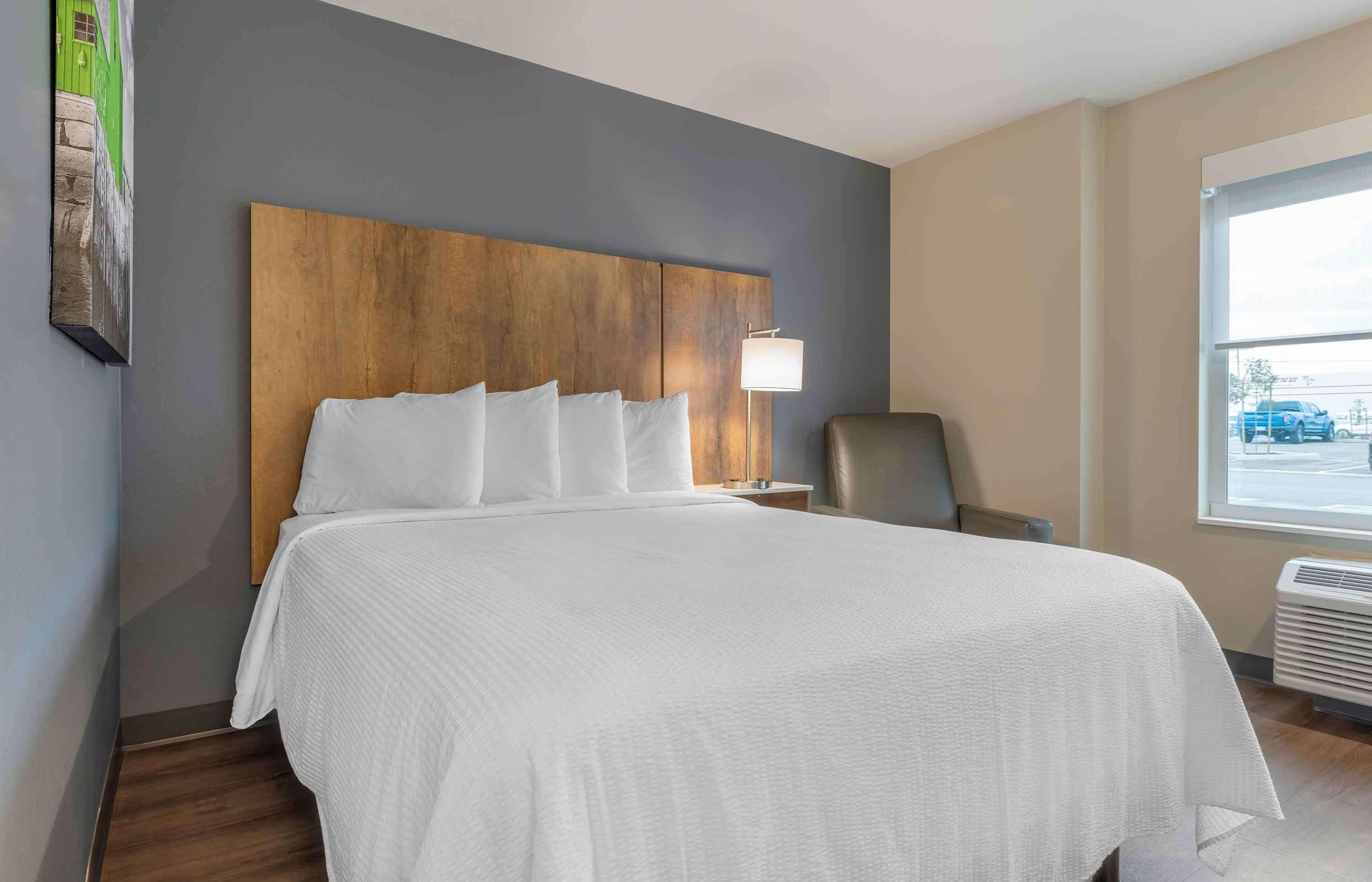 A hotel room with a large bed, a wooden headboard, a nightstand with a lamp, a chair, and a window with a view of the outside.