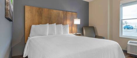 A hotel room with a large bed, a wooden headboard, a nightstand with a lamp, a chair, and a window with a view of the outside.