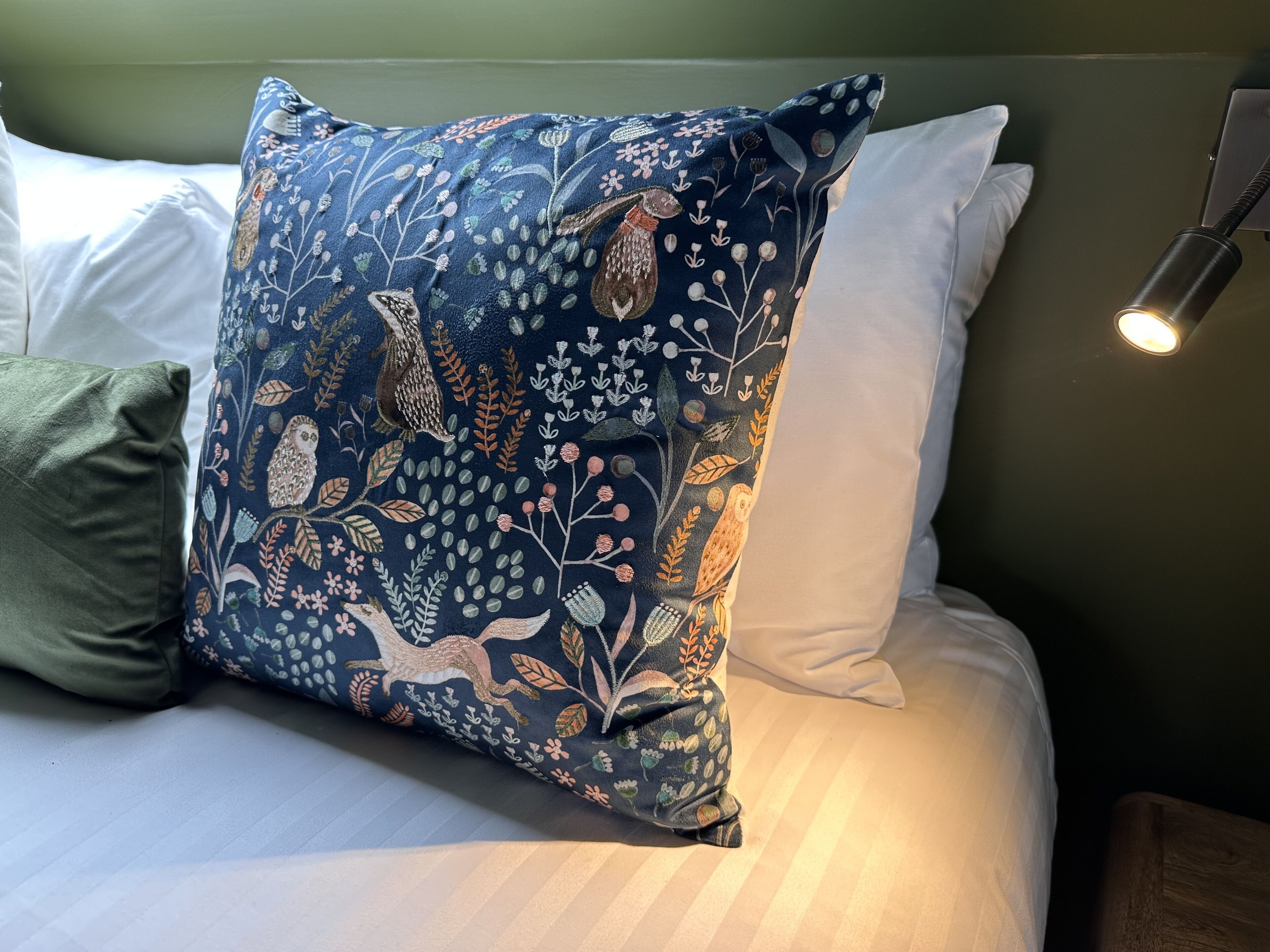 A bed with a decorative pillow featuring a bird and floral pattern, a green pillow, and a bedside lamp.