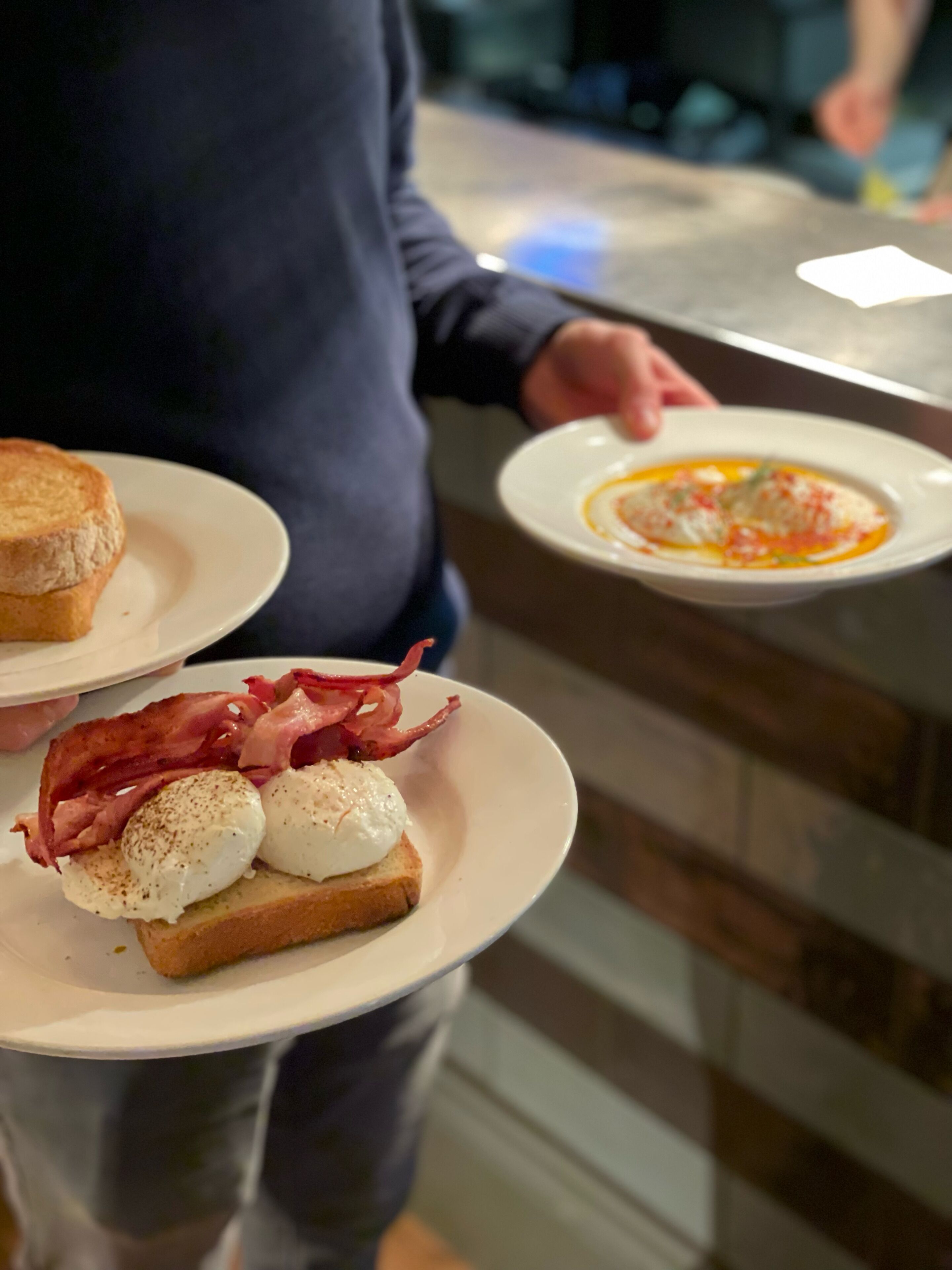 A person holding two plates, one with toast and eggs, and another with a soup dish.
