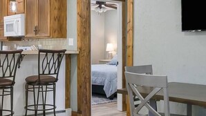 Une cuisine compacte avec des meubles en bois et un four à micro-ondes, un coin repas avec deux tabourets de bar et une table, et une chambre à coucher visible à travers une porte ouverte.