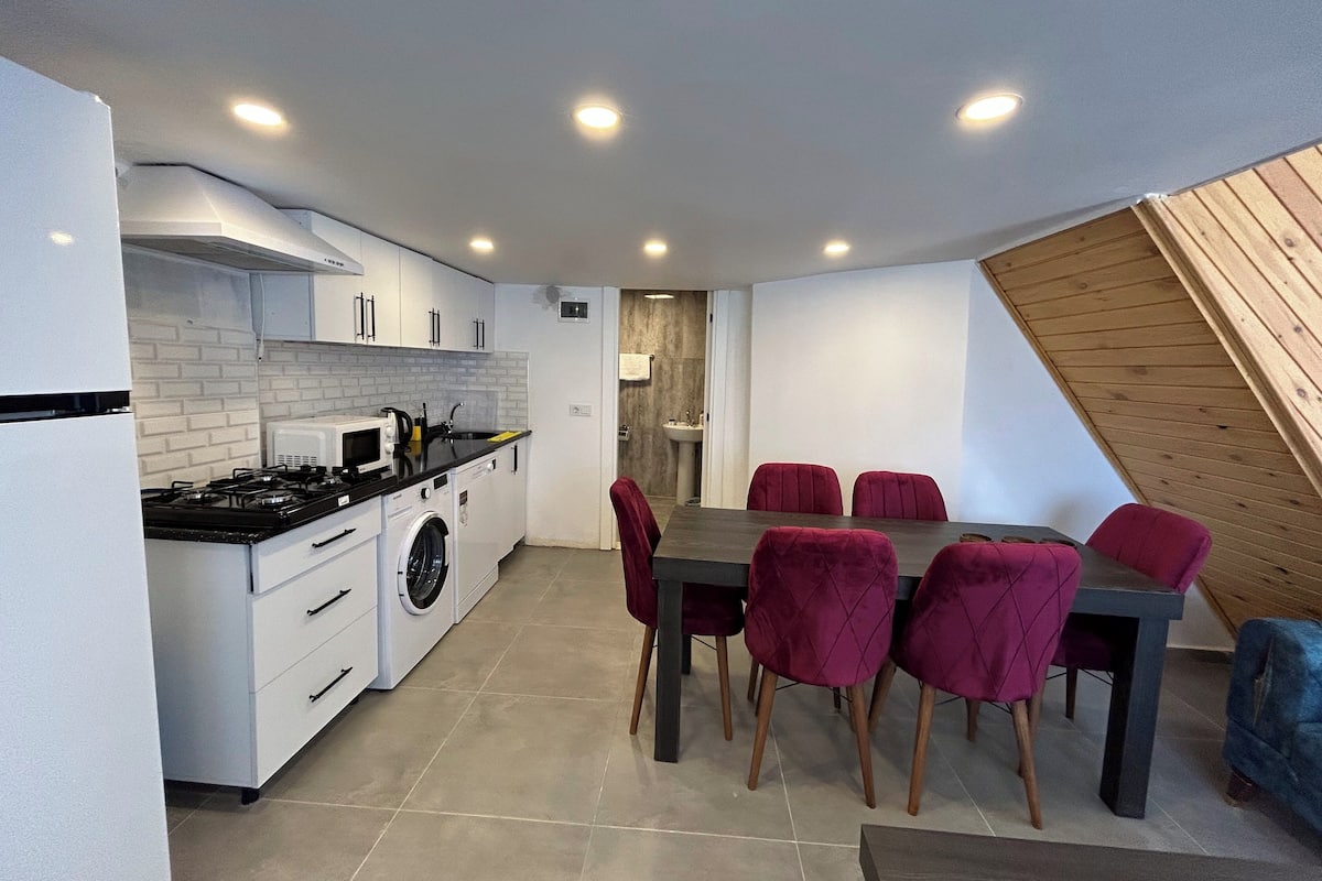 A compact kitchen with white cabinets, a stove, and a washing machine. There is a dining table with six chairs and a wooden sloped ceiling.