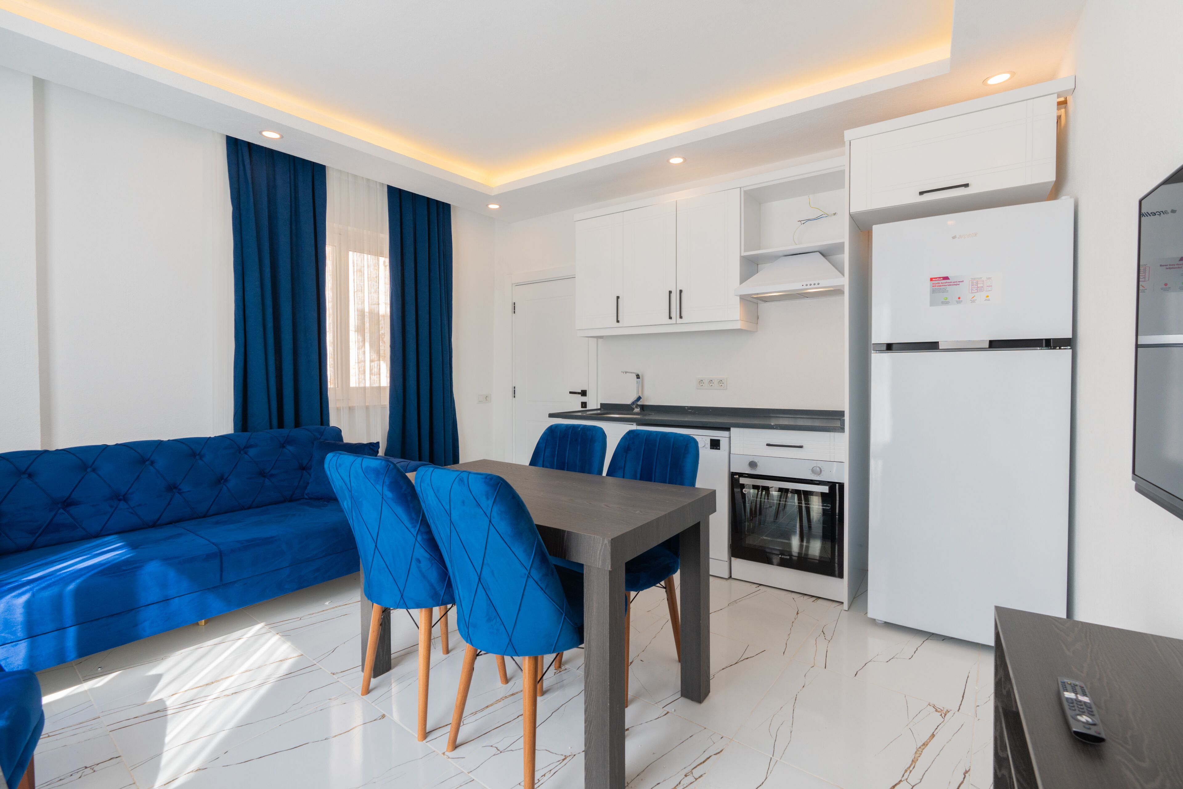Una sala moderna con un sofá azul, una mesa de comedor con sillas azules, una zona de cocina con armarios blancos, un horno empotrado y un refrigerador.