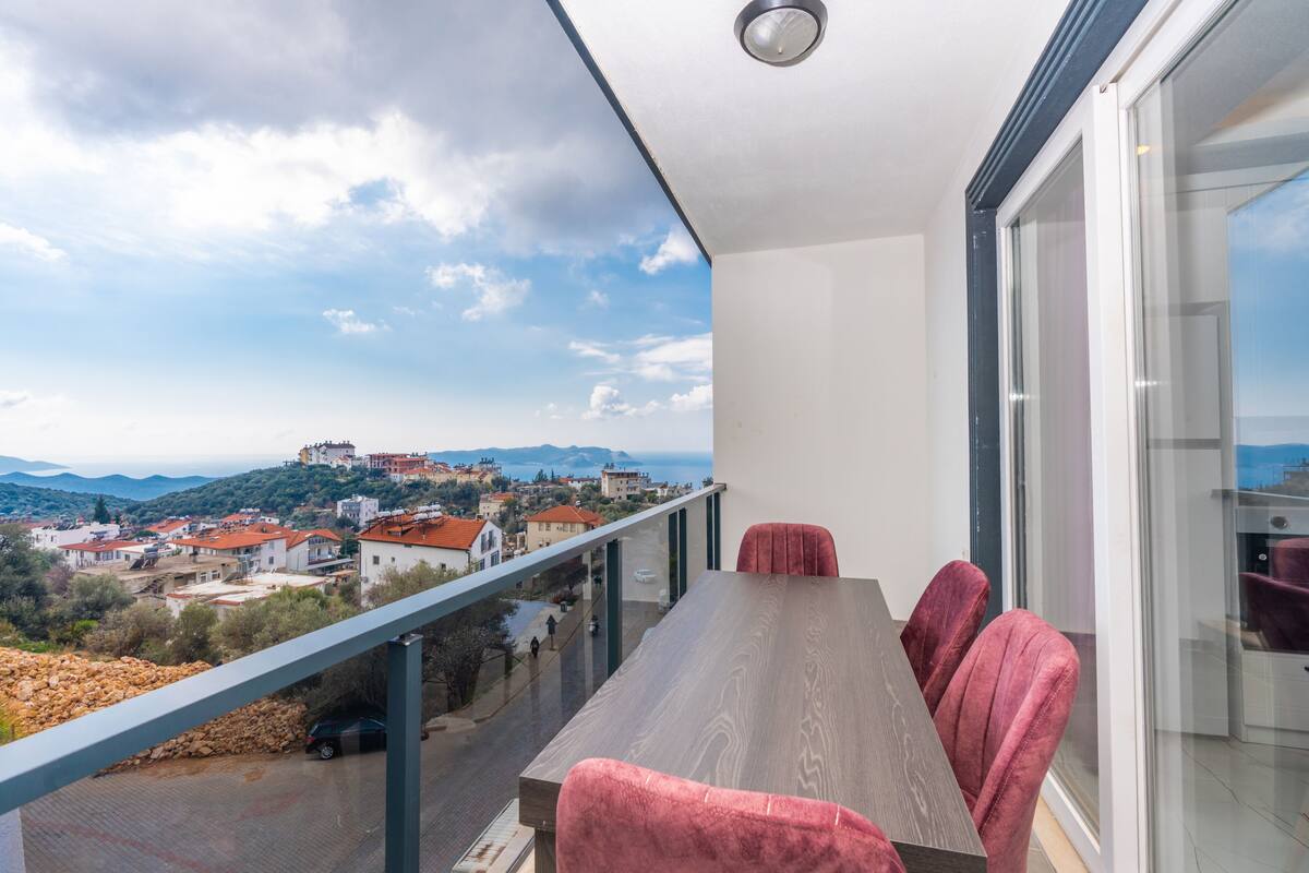 A balcony with a wooden table and red chairs, overlooking a coastal town with buildings and mountains in the distance.