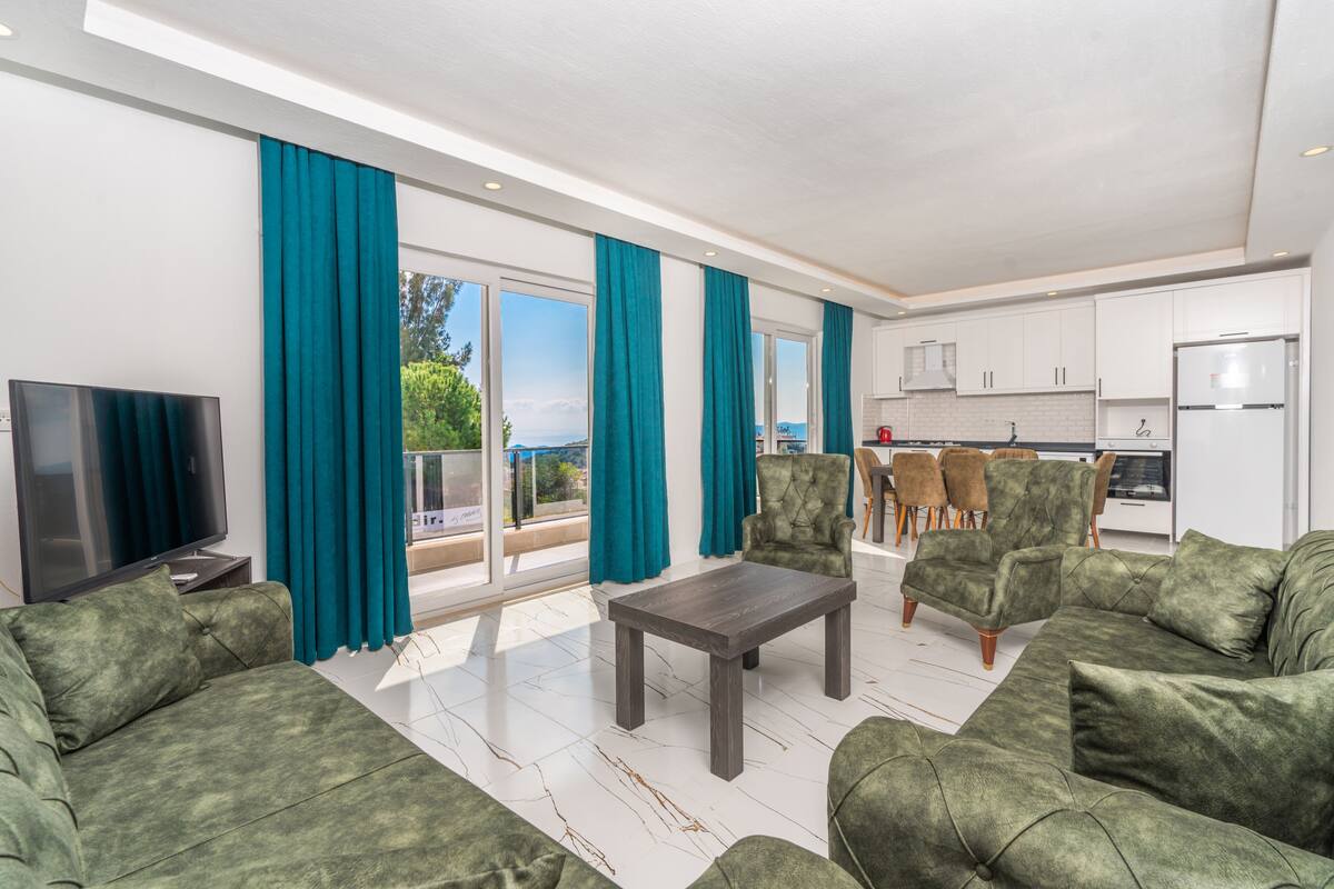 A modern living room with green sofas, a coffee table, and a TV. There is a balcony with a view of the sea.