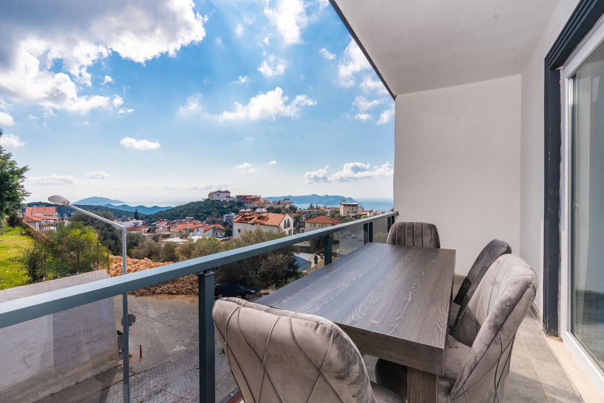 A balcony with a dining table and chairs, overlooking a town with red-roofed buildings and a clear sky.