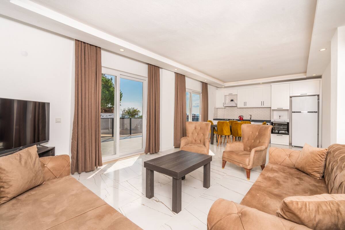 A modern living room with a sofa, a coffee table, and a TV. There is a kitchen in the background with a dining area.