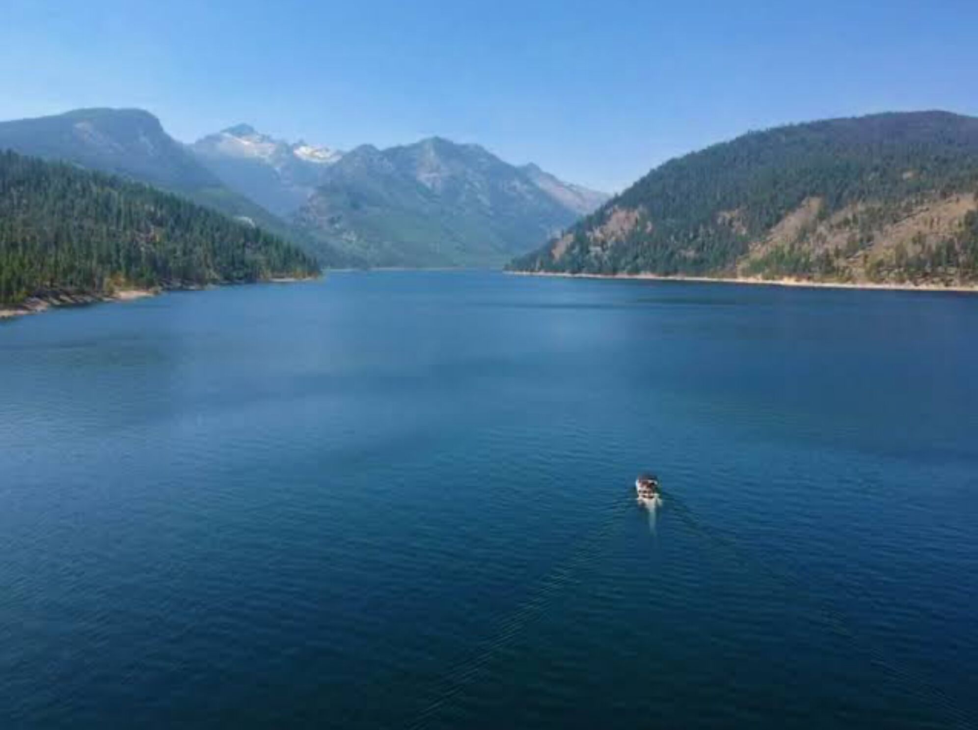 A calm lake with a small boat in the center, surrounded by forested mountains under a clear sky.