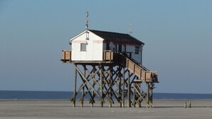 A wooden pier structure with a small building on top, likely a lifeguard station, on a sandy beach with the ocean in the background.
