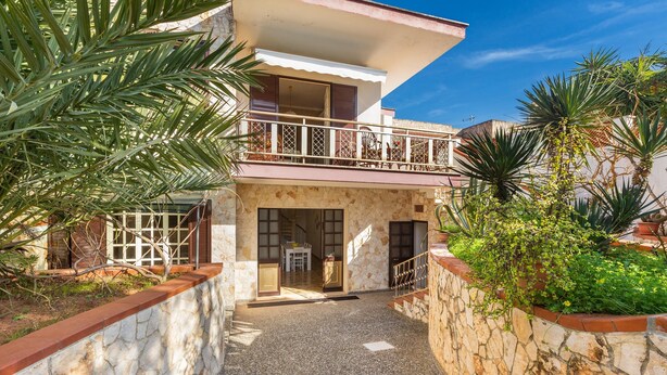 A two-story house with a balcony, stone walls, and tropical plants.