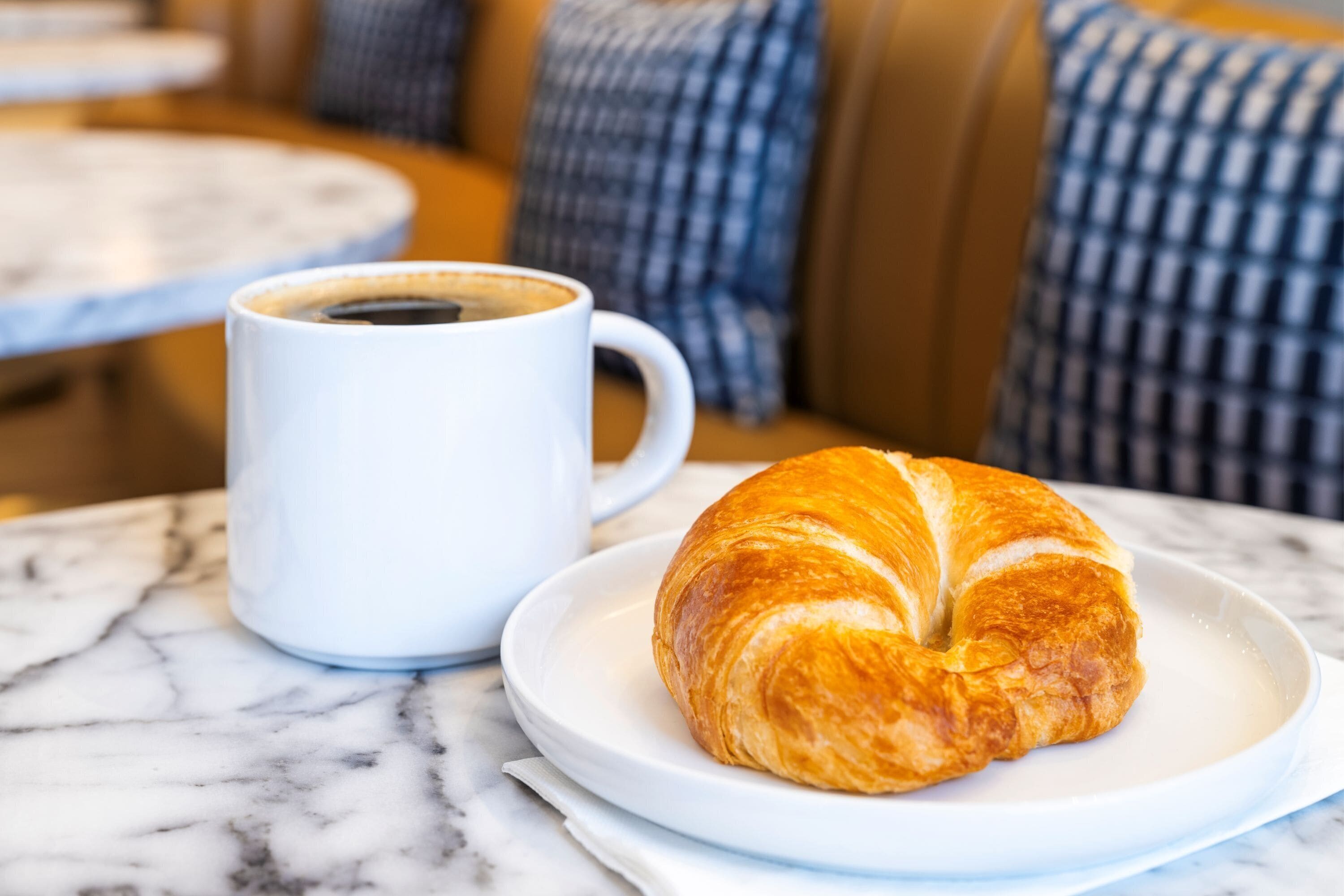 Un croissant sobre un plato blanco, junto a una taza de café en una mesa de mármol.