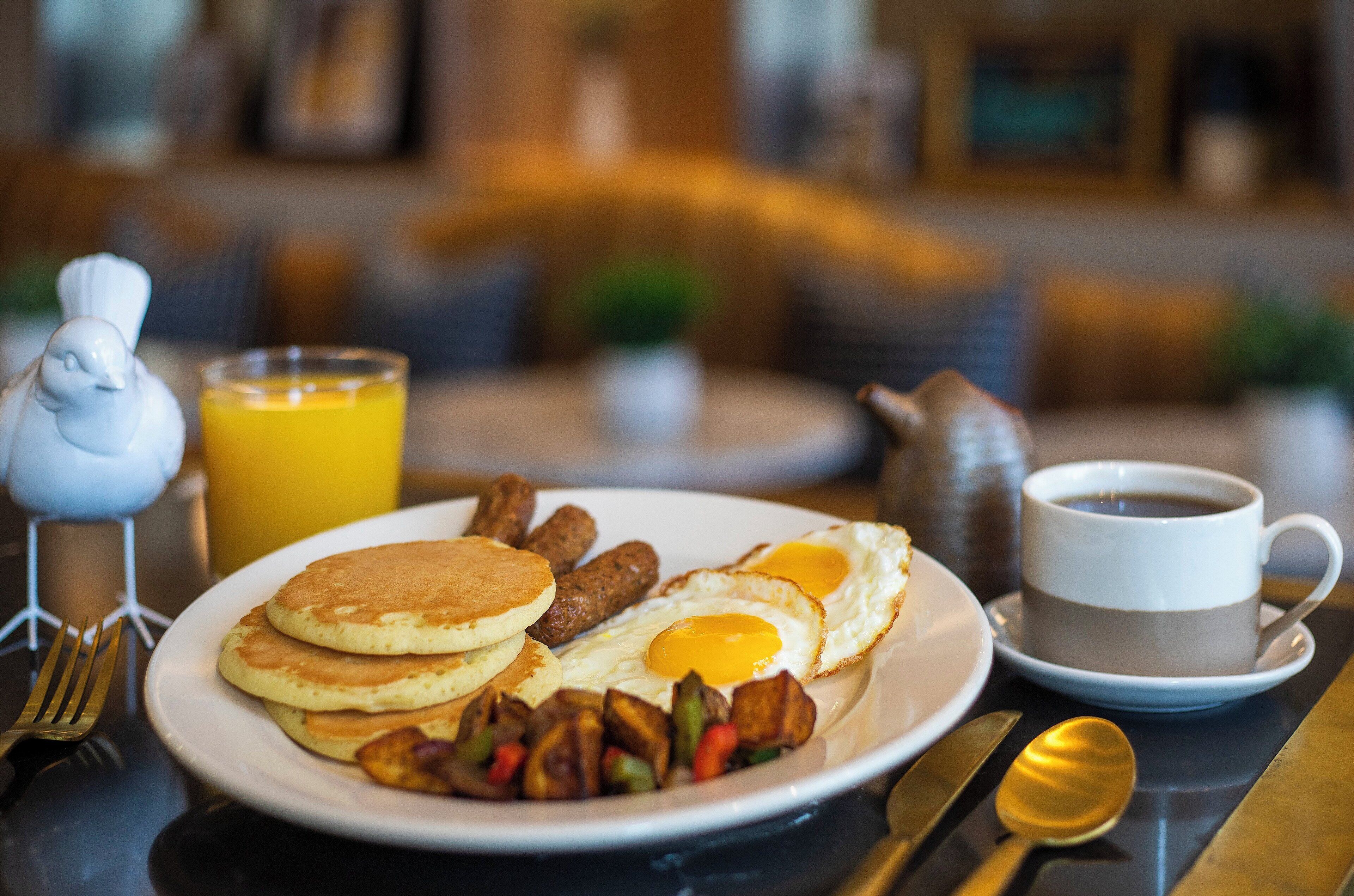 Un plat de petitdejuni amb crepes, ous, salsitxes i verdures, acompanyat d'un got de suc d'aranja i una tassa de cafè.