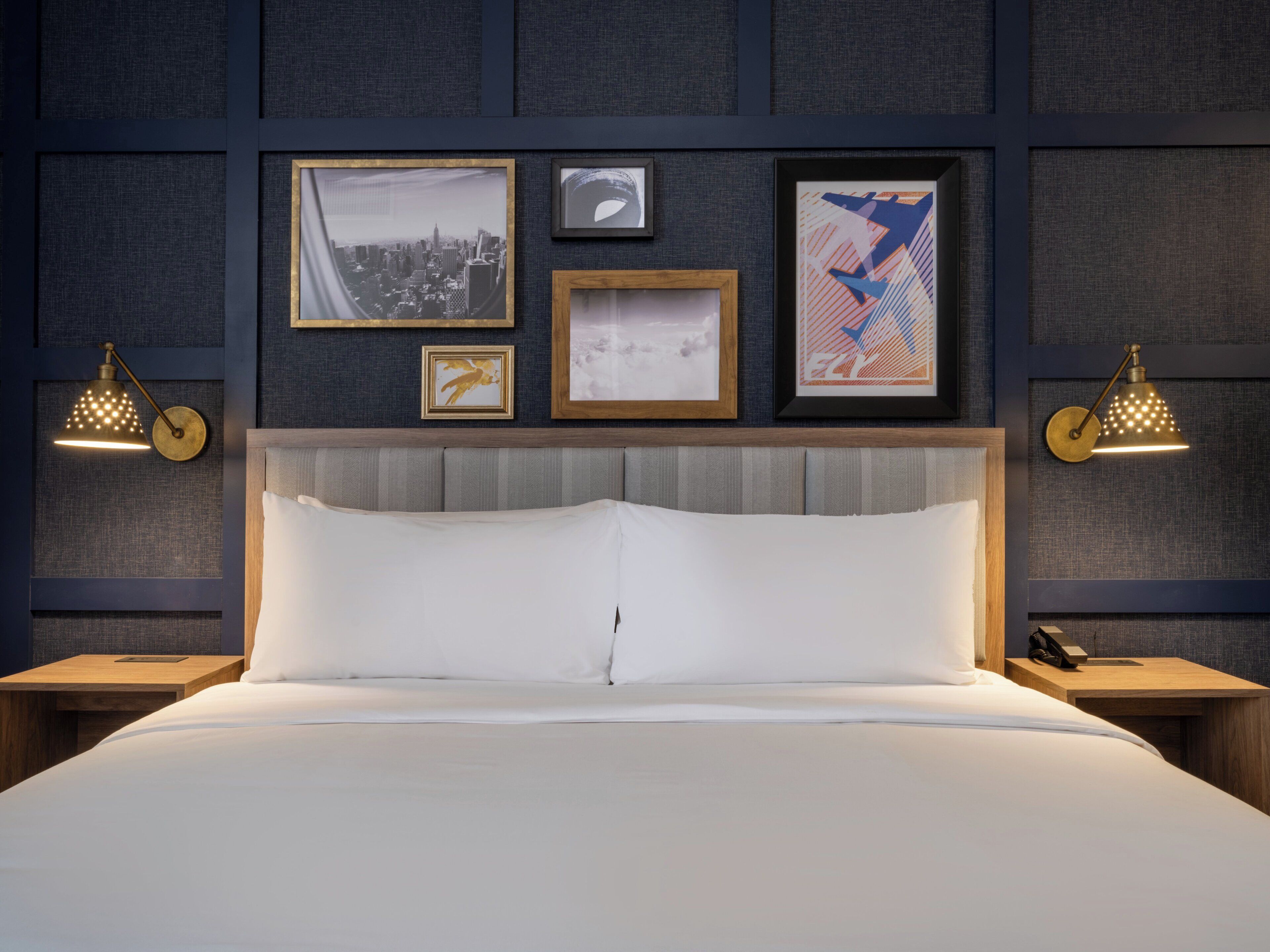 A hotel room with a large bed, a headboard, and framed pictures on the wall. There are two bedside tables with lamps.