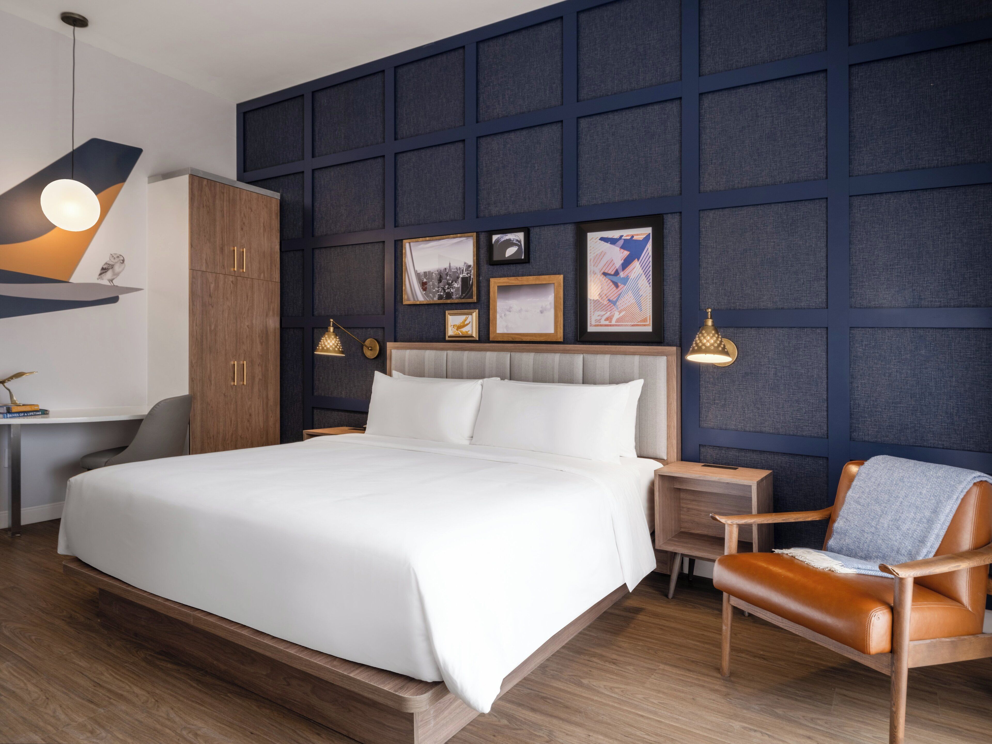 Una habitación de hotel moderna con una cama grande, cabecera de madera y una pared de acento azul oscuro. Hay dos mesitas de noche con lámparas, un armario de madera y un sillón de cuero.