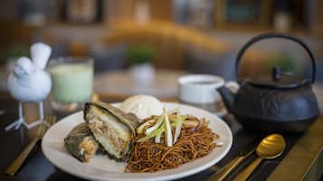 A plate of food with noodles and a wrapped item, a teapot, and a cup on a table.