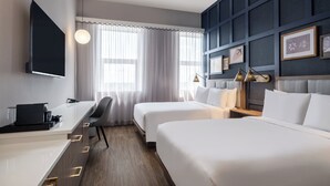 Une chambre d’hôtel avec deux lits, un bureau, un téléviseur à écran plat et une grande fenêtre avec des rideaux légers.