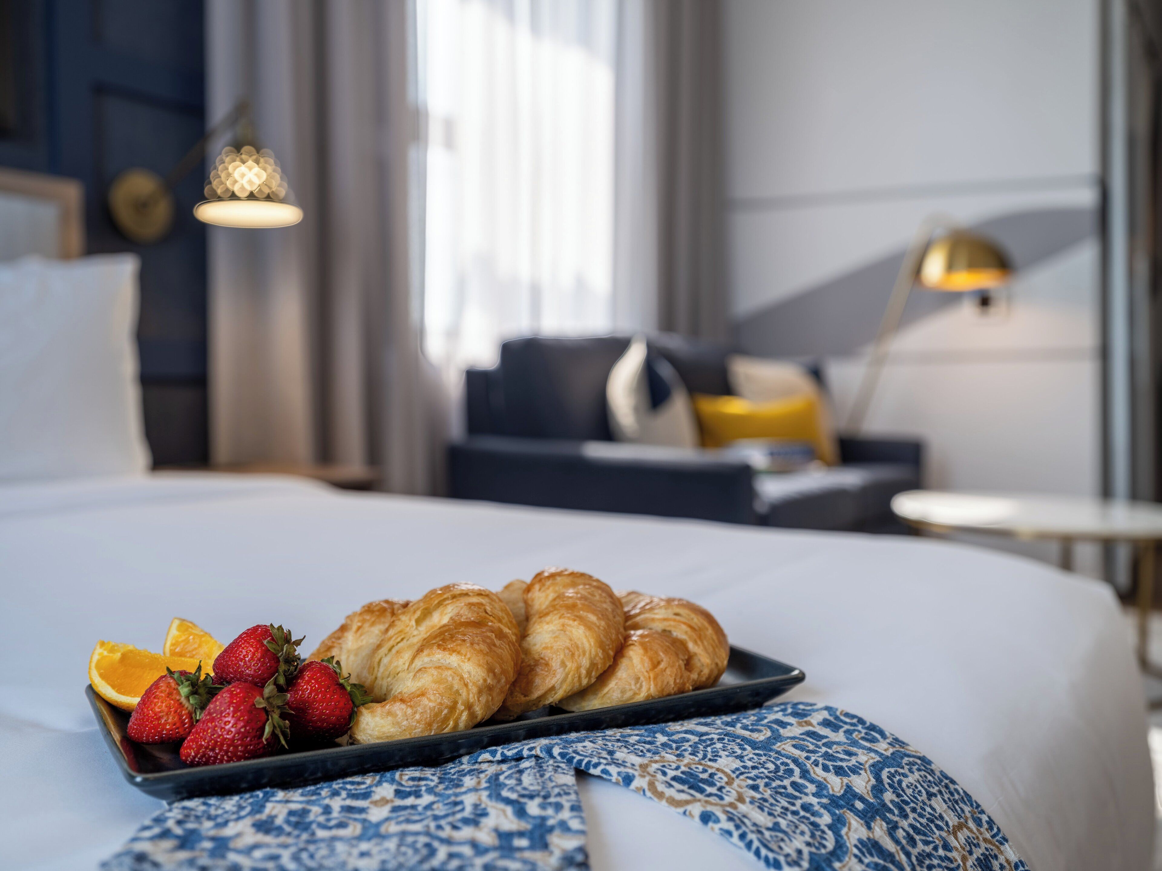 A neatly made bed with a tray of croissants and strawberries, a patterned throw, and a modern room with a sofa and lamps.