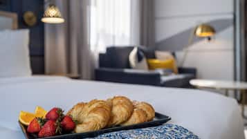 A neatly made bed with a tray of croissants and strawberries, a patterned throw, and a modern room with a sofa and lamps.