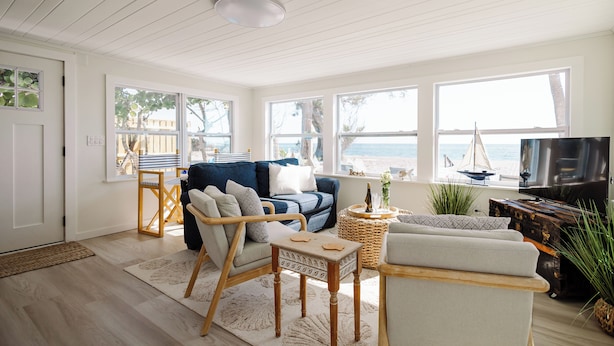 A spacious living room with a blue sofa, wooden chairs, a TV, and large windows overlooking the sea. - "Lost Loon" Oceanfront Cottage by Roxy Rentals (Englewood)