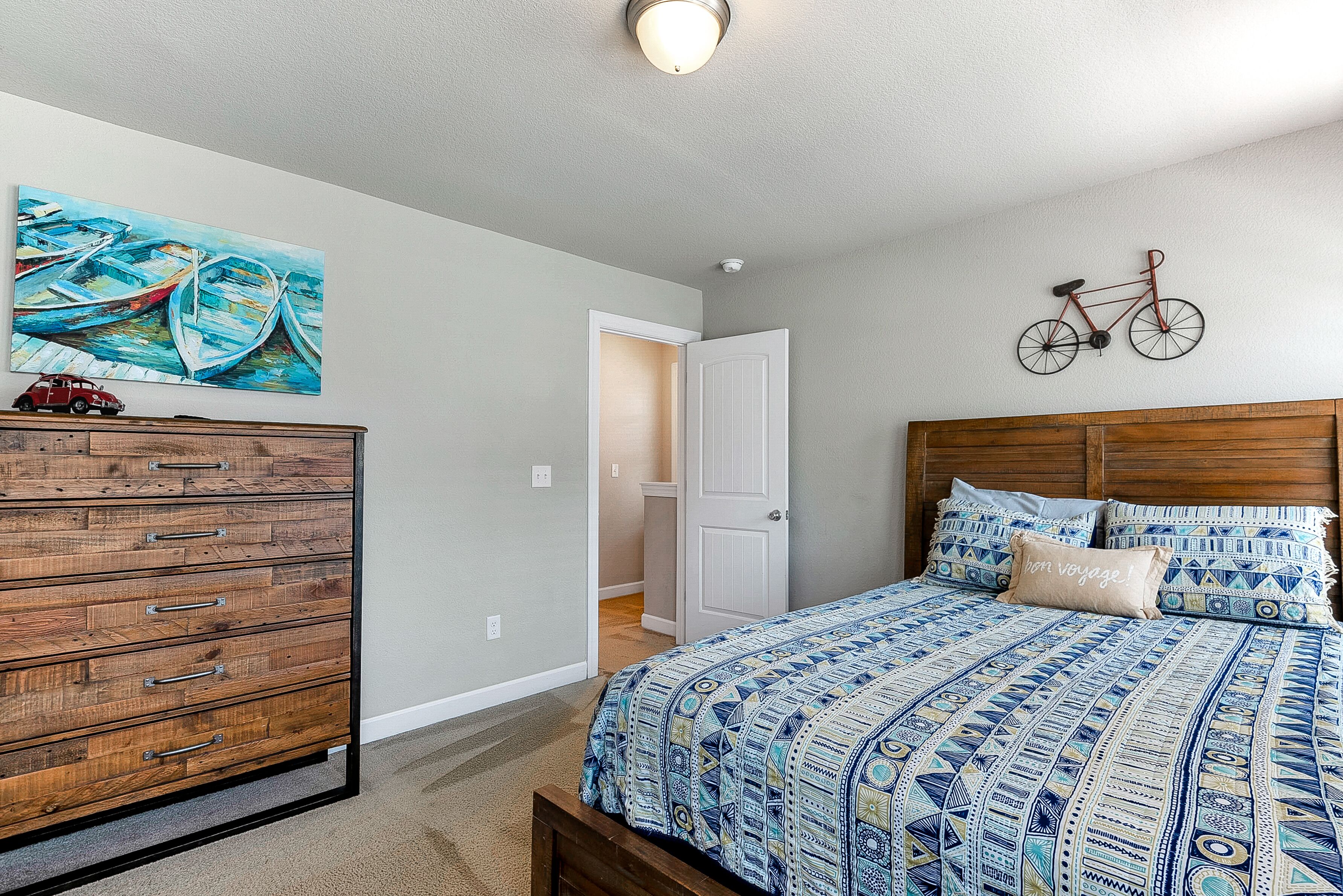 A bedroom with a wooden dresser, a bed with patterned bedding, a bicycle wall decoration, and a painting of boats.
