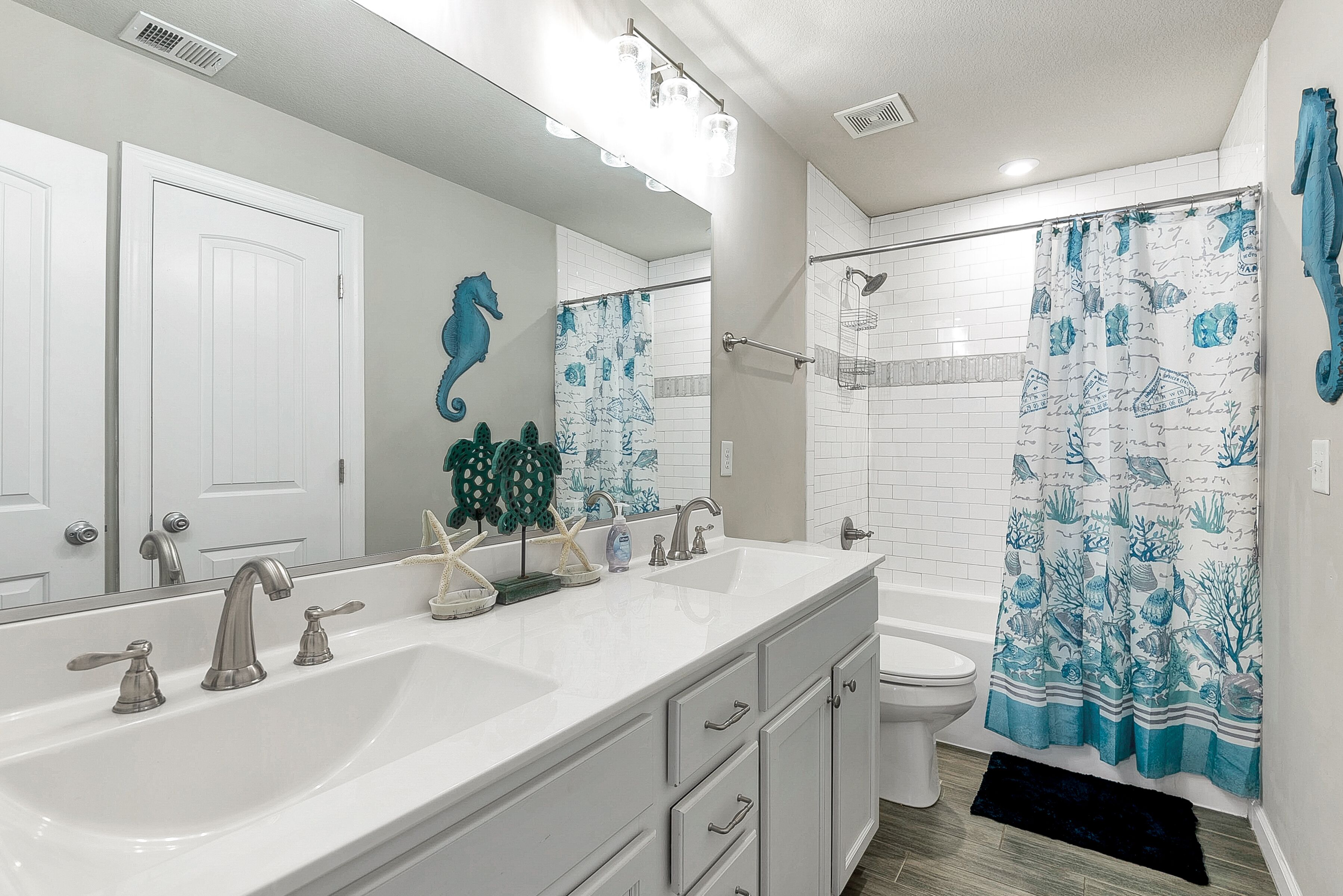 Une salle de bain avec un lavabo blanc, deux robinets, un miroir, un rideau de douche orné de motifs représentant des hippocampes et des toilettes blanches.