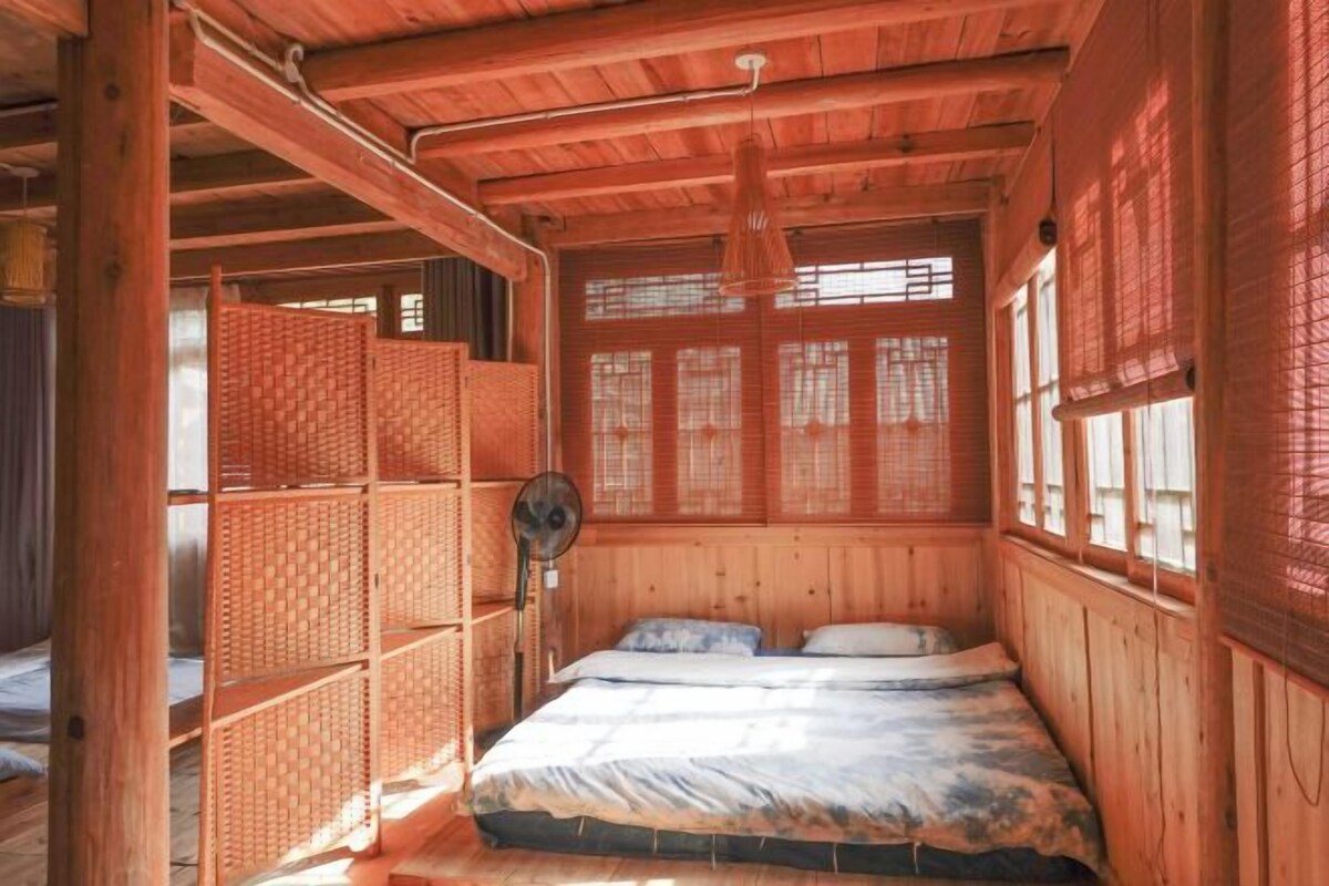Quarto de madeira com uma cama, um ventilador e uma janela com persianas.
