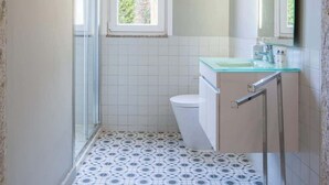 Casa de banho com chão estampado, sanita, lavatório com bancada de vidro e espelho.