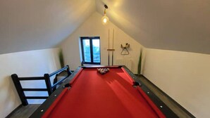 A pool table with a red felt surface, a white cue ball, and a few scattered billiard balls. There is a window with a view of buildings outside, a black metal bench, and a light fixture on the ceiling. - Le Bill'ppart Easy Life (Bourgoin-Jallieu)