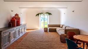 A spacious living room with a large window, a sofa, a dining table, and a cabinet. - Cenoura-Brava Villas & Suites. (Évora)