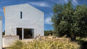 A modern white building with a large glass door and windows, surrounded by natural vegetation and a clear blue sky. - Cenoura-Brava Villas & Suites. (Évora)