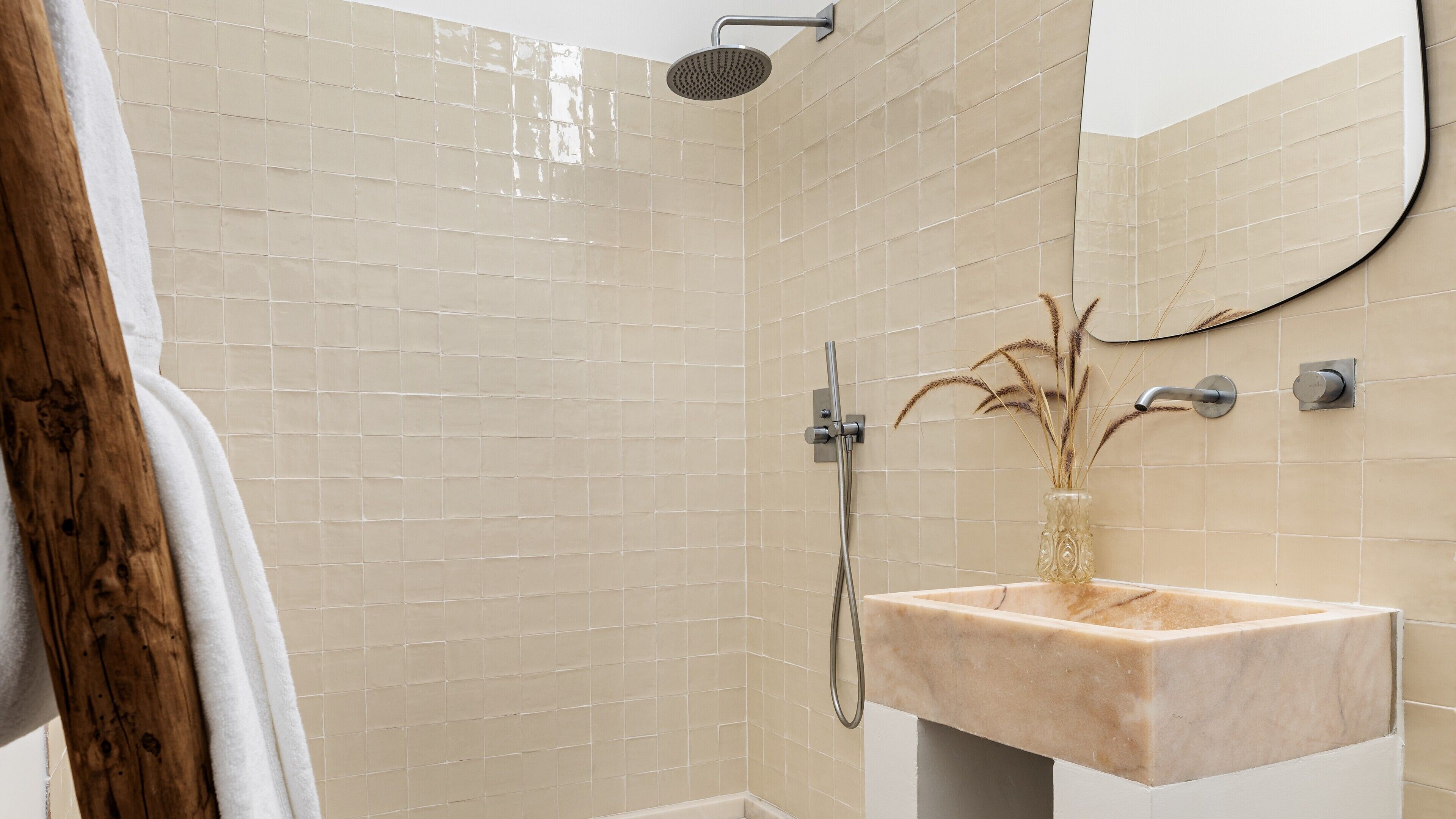 Um banheiro com paredes revestidas de azulejos bege, uma pia de mármore, chuveiro manual e um espelho.