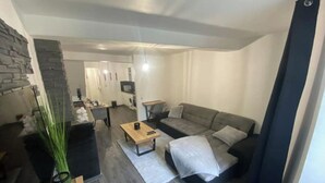 A modern living room with a grey sofa, a coffee table, and a TV. There is a kitchen area in the background with a washing machine. - Le Ciné'ppart Easy Life (Bourgoin-Jallieu)