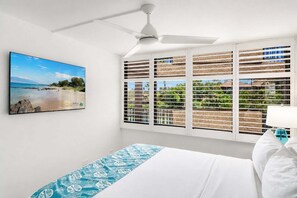 Habitación de hotel con cama, ventilador de techo, ventana con persianas y un cuadro con una playa.