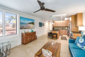 A spacious living area with a large window, a ceiling fan, a wooden dining table, a blue sofa, and a kitchen with wooden cabinets and a countertop.