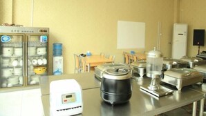 Un coin cuisine équipé d’un réfrigérateur, d’une machine à café, d’une bouilloire et d’un four à micro-ondes. On aperçoit des chaises et une table au fond.