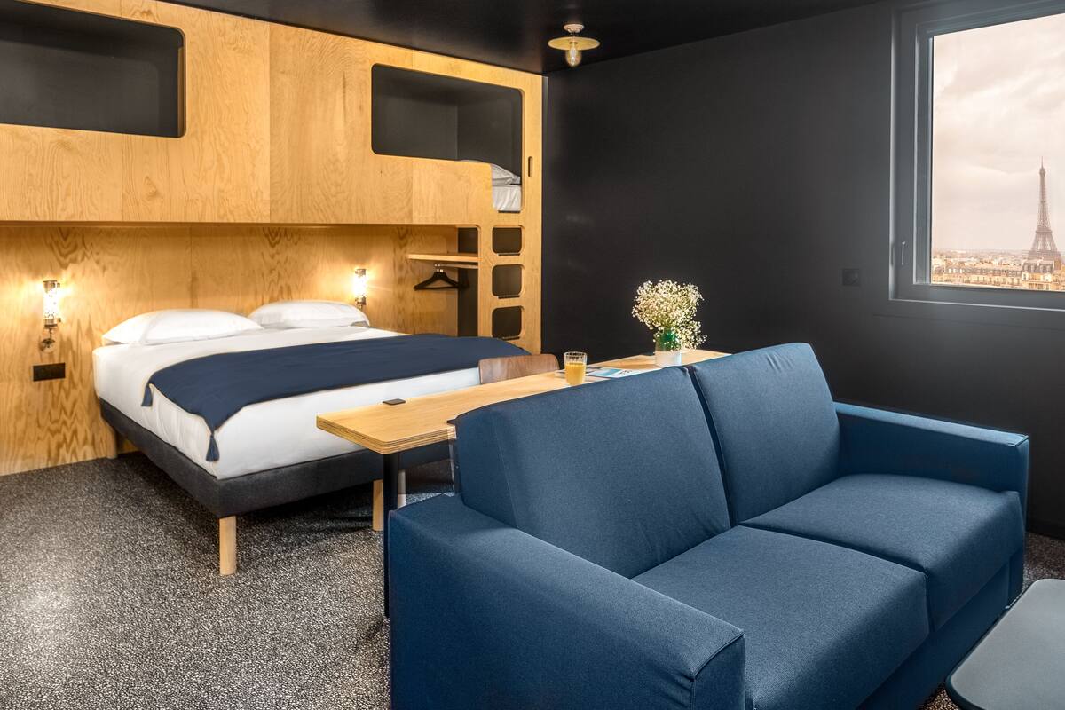 Ein modernes Hotelzimmer mit Bett, blauem Sofa, kleinem Tisch und Fenster mit Blick auf die Stadt.