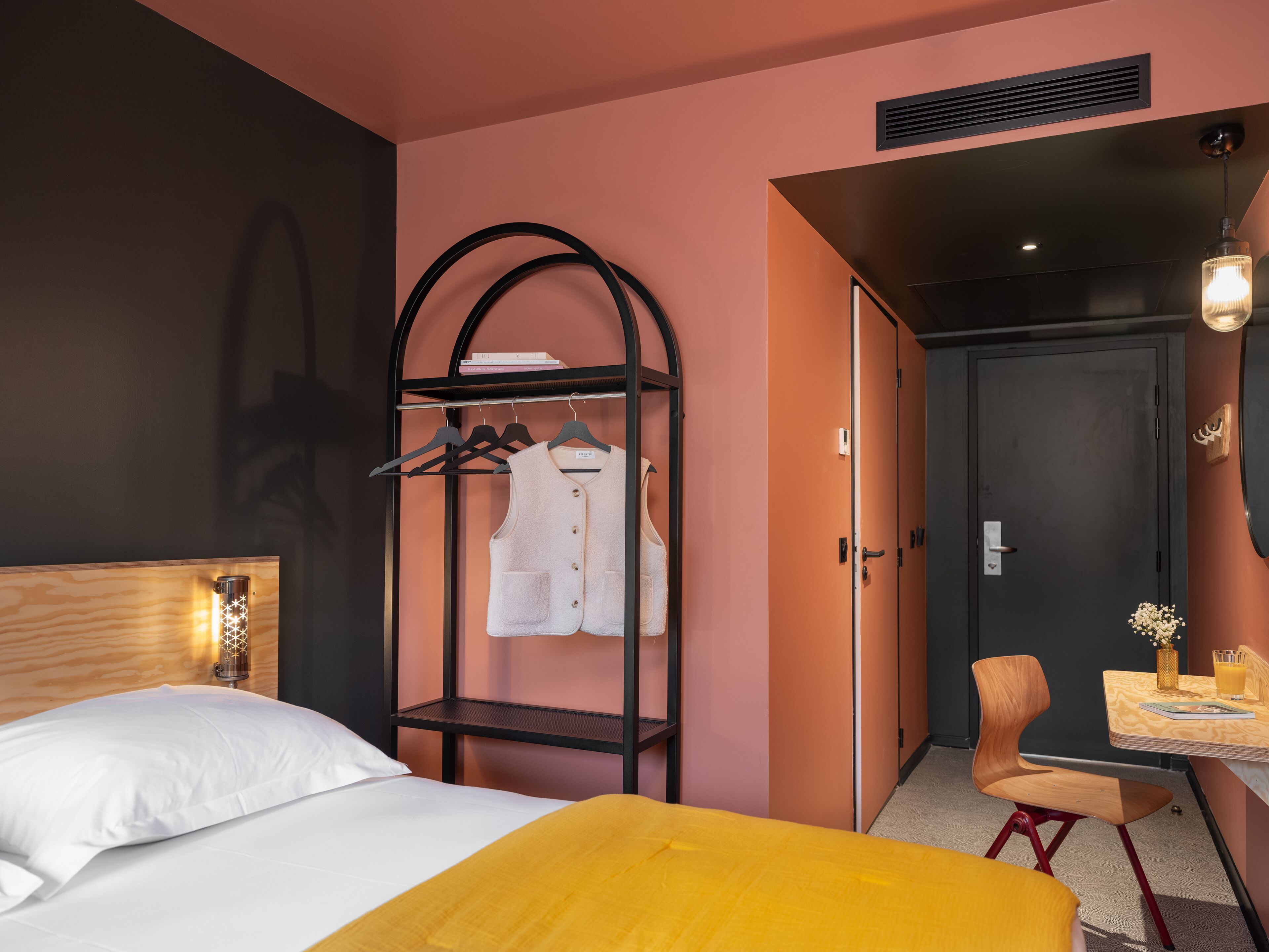 A hotel room with a bed, a wooden nightstand, a hanging coat rack, a desk with a chair, and a door.