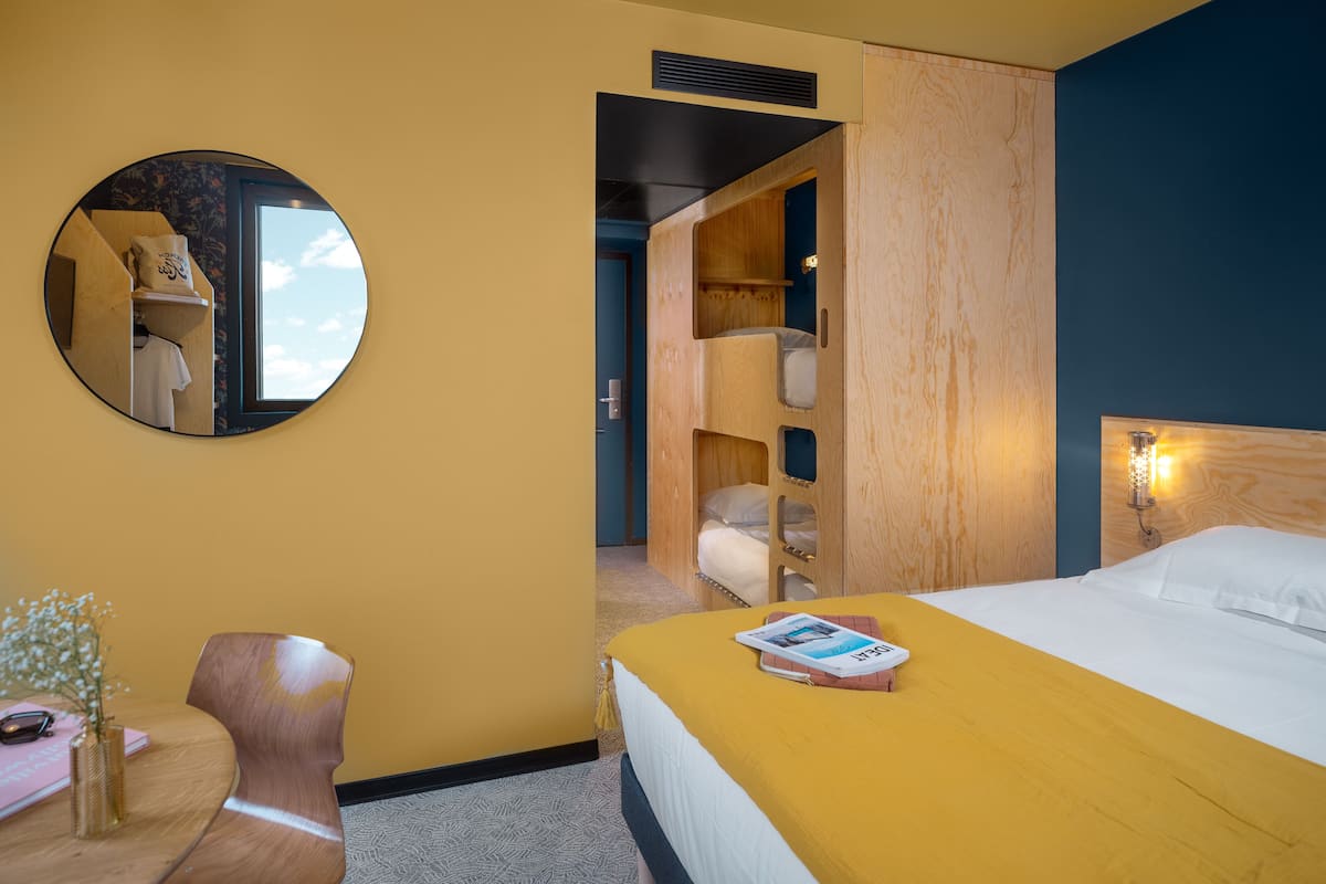 Ein Hotelzimmer mit einem Bett, einem runden Spiegel, einem Holzschrank und einem kleinen Tisch mit Vase und Buch.