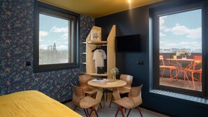 Habitación de hotel con cama, mesa redonda con sillas, televisor montado en la pared y balcón con sillas y mesa de color naranja.