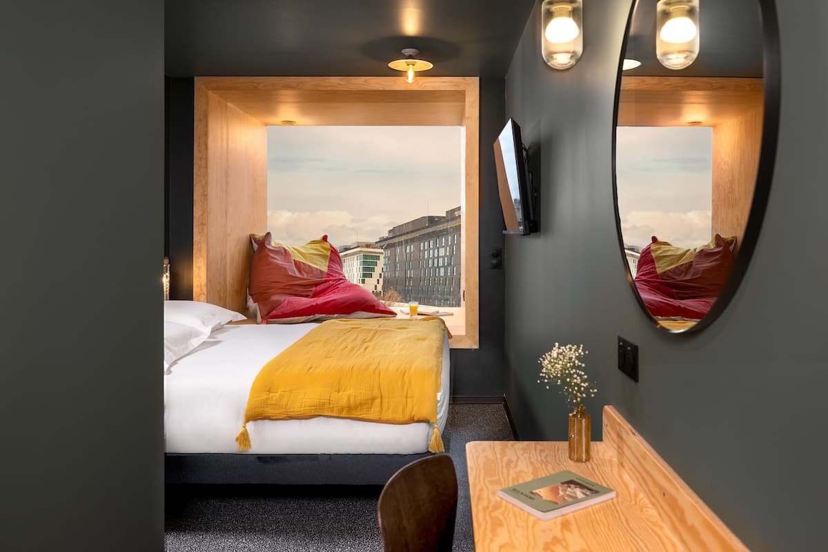 Ein modernes Hotelzimmer mit einem großen Fenster, einem Bett mit gelber Tagesdecke, einem Holztisch mit einer Vase und einem Spiegel, der den Raum reflektiert.
