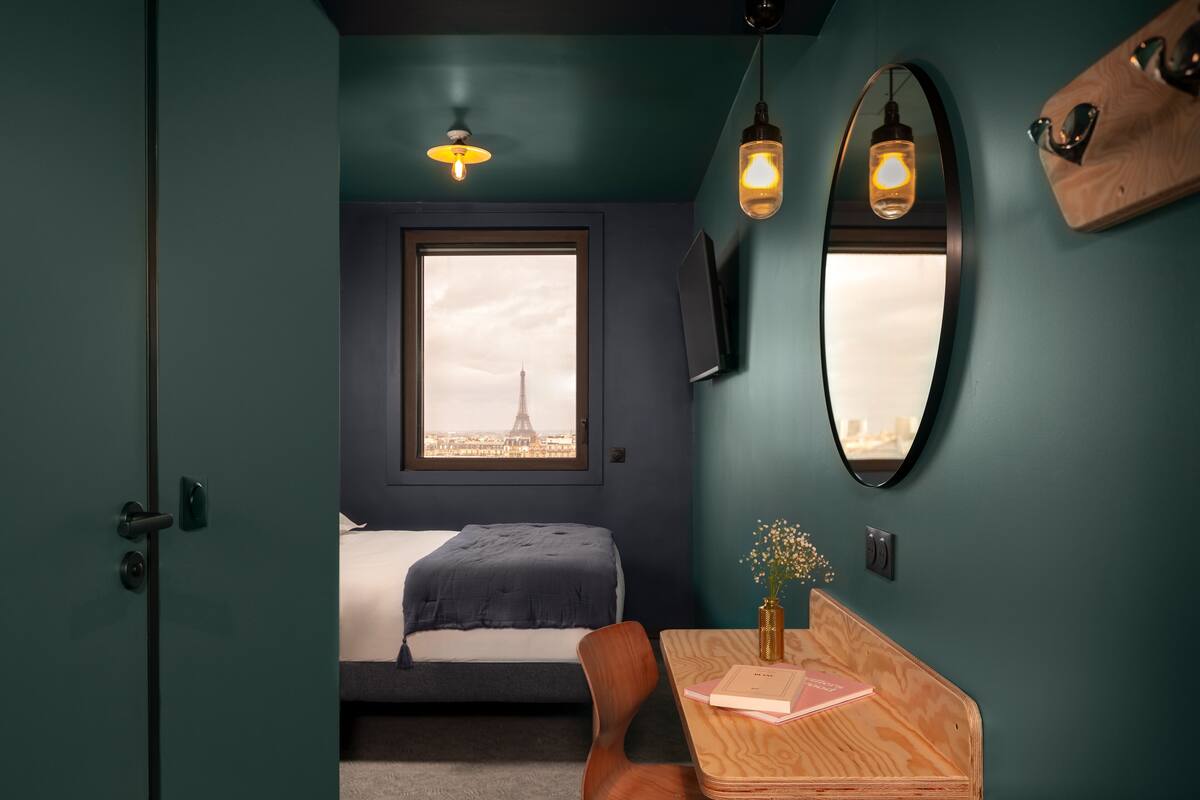 Ein Schlafzimmer mit Blick auf den Eiffelturm, ein Bett, ein Schreibtisch mit Büchern und ein Spiegel an der Wand.