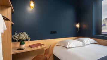 A modern bedroom with a dark blue wall, a wooden desk with a chair, a bedside table with a vase of flowers, a bed with white pillows, and a hanging light fixture.